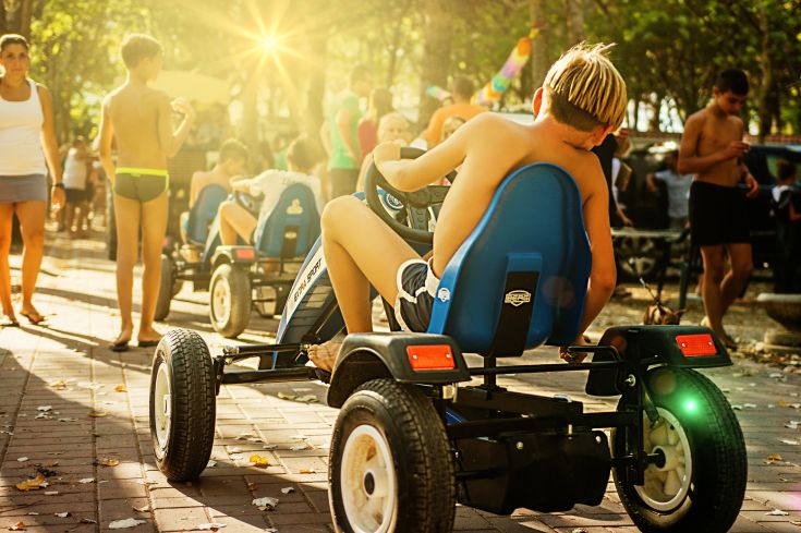 Un bambino conduce un go-kart in un parco illuminato dal sole, in mezzo ad altre persone che si divertono.
