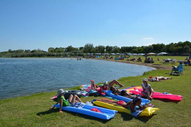 Un pomeriggio di sole in riva al lago, con persone distese su materassini colorati sull'erba.