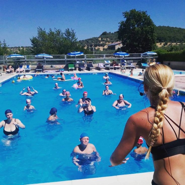 Lezione di fitness in piscina con partecipanti attivi, sotto un cielo sereno e soleggiato, circondati da vegetazione.