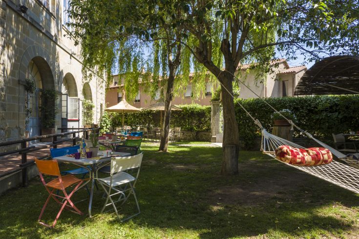 Ein Garten mit bunten Tischen, einer Schaukel und Bäumen, die an einem sonnigen Tag Schatten spenden.