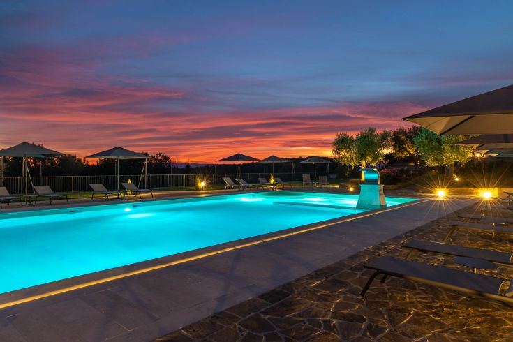 A swimming pool in greenery, surrounded by trees, with the sun setting on the horizon.