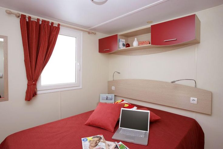 Simple bedroom with a double bed, functional furnishings, and large windows providing natural light.