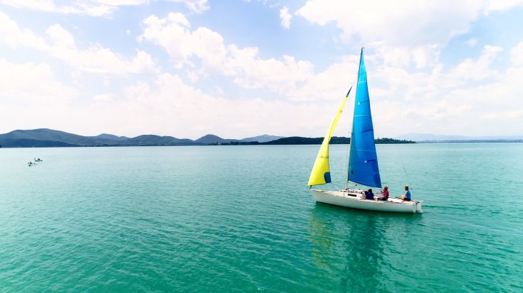 A brightly colored sailboat glides over the calm waters of a lake, with mountains in the background.