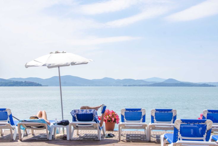 A serene area with blue sun loungers and umbrellas, overlooking a lake and distant hills.