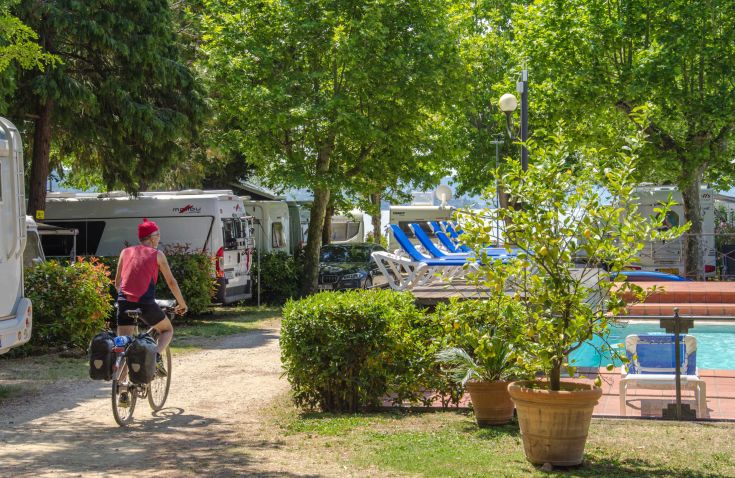 A cyclist rides in a green area, near a campground and a pool, surrounded by nature.