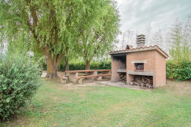 An outdoor barbecue area with a brick grill, surrounded by greenery, trees, and plants.