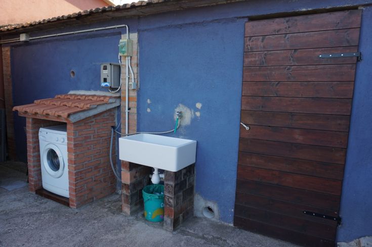 Area esterna con lavatrice e lavello, dotata di una porta in legno su una parete blu.