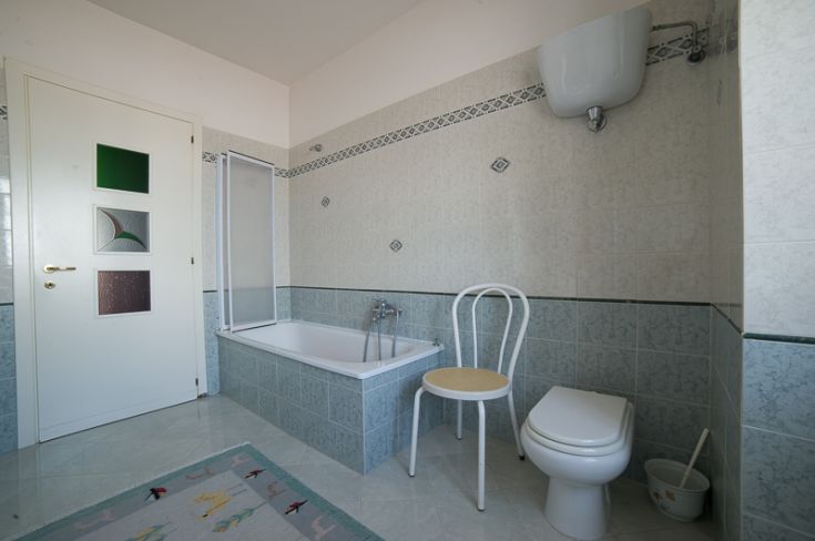 Un bagno spazioso e ben illuminato, dotato di vasca, wc e una sedia in legno chiaro.