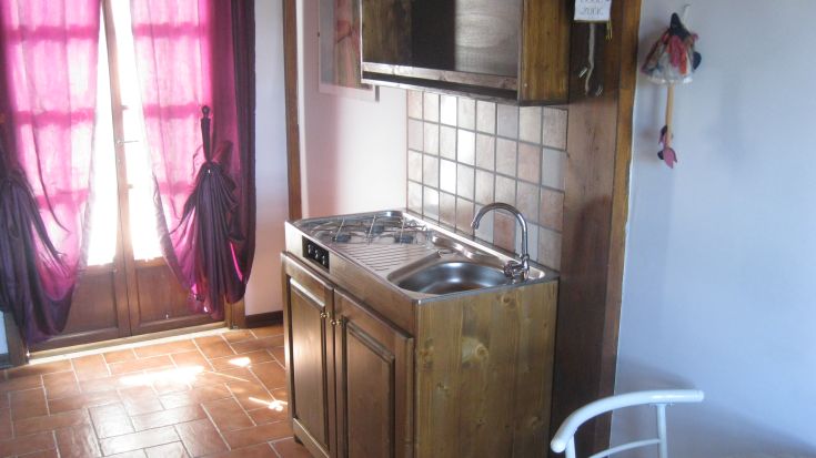 Cucina semplice con mobili in legno e tende color bordeaux, illuminata dalla luce naturale.