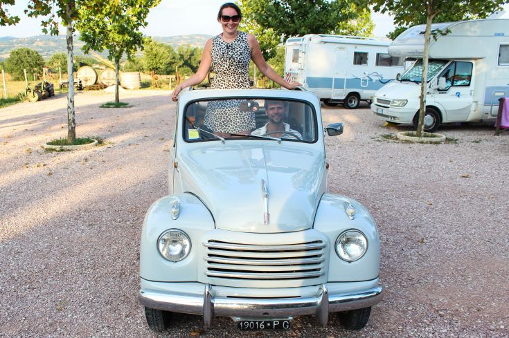 Una coppia sorridente si trova accanto a un'auto d'epoca in un contesto rurale tranquillo.