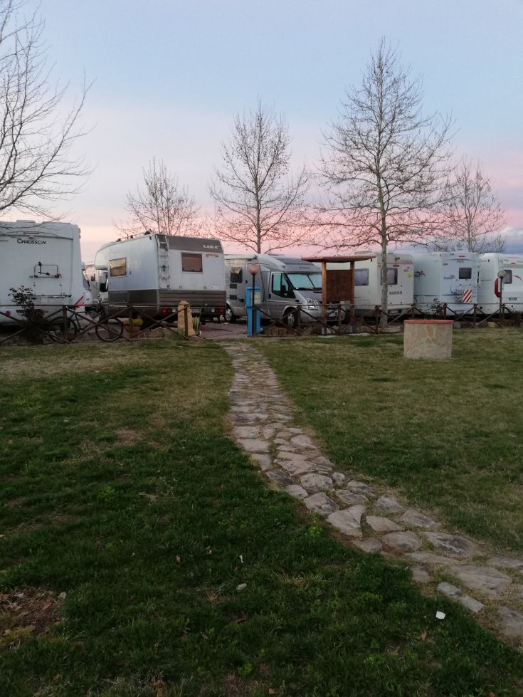 Area di campeggio silenziosa con camper, immersa nel verde e un cielo chiaro al tramonto.