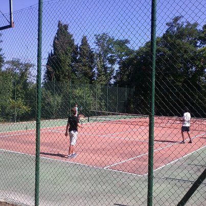 Due giocatori si sfidano su un campo da tennis circondato dalla vegetazione. L'ambiente è sereno e verde.