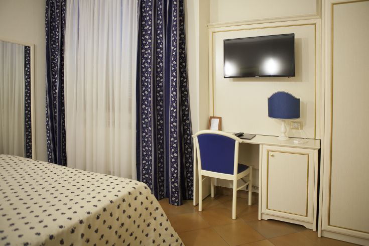 Camera semplice con arredi funzionali, dotata di televisore e finestra decorata da tendaggi.