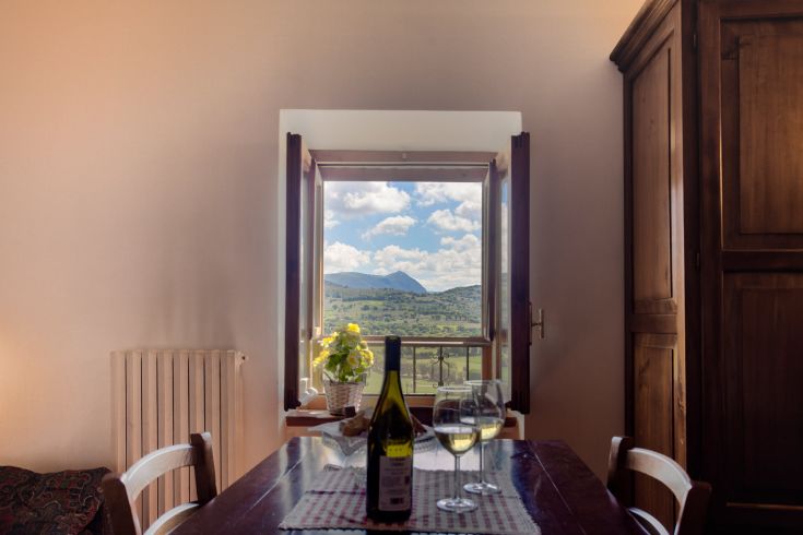 Scena da una finestra che mostra un classico paesaggio montano, con un tavolo apparecchiato e una bottiglia di vino.