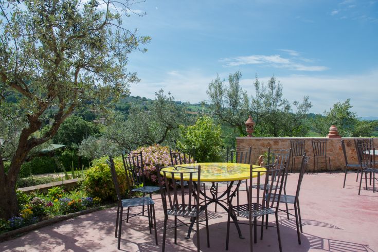Una terrazza immersa nella natura, con un tavolo giallo e sedie. Circondata da ulivi e piante fiorite.