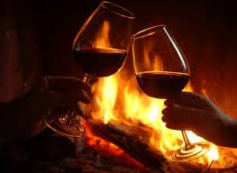 Due bicchieri di vino rosso si trovano accanto a un camino acceso, offrendo un'atmosfera calda e conviviale.