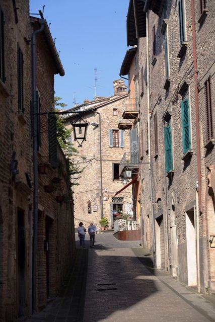 Une ruelle avec des bâtiments en pierre et des fenêtres vertes, où deux personnes se promènent tranquillement.