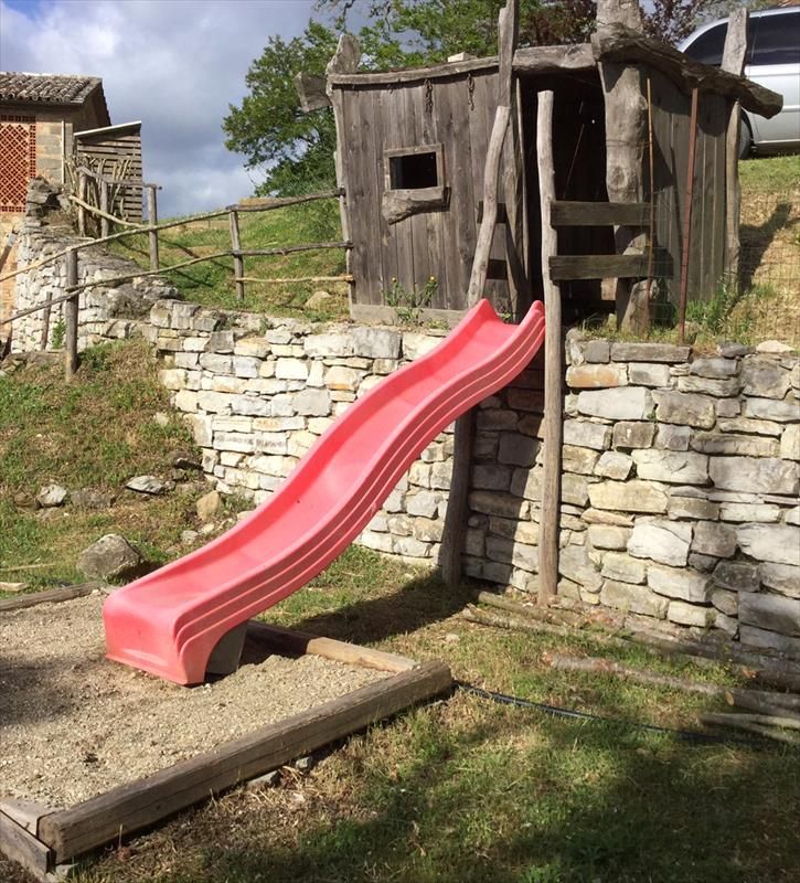 A pink slide in a playground, surrounded by green trees and bushes. A wooden structure is visible in the background.