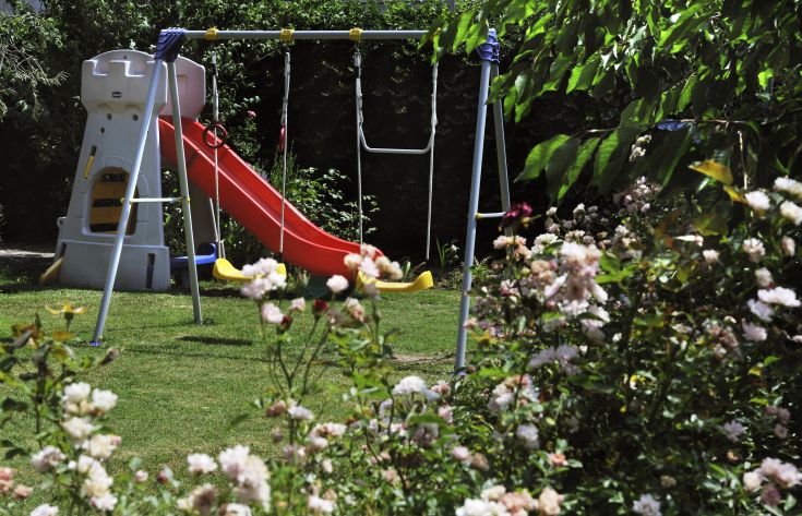 Ein Spielbereich im Garten mit einer Rutsche und Schaukeln, umgeben von verschiedenen Pflanzen und Blumen.