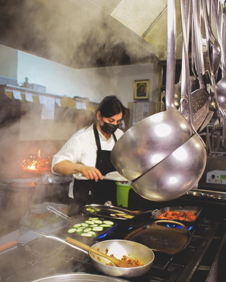 Ein Koch bereitet Gerichte in einer traditionellen Küche zu, mit hängenden Utensilien und frischen Zutaten auf dem Herd.