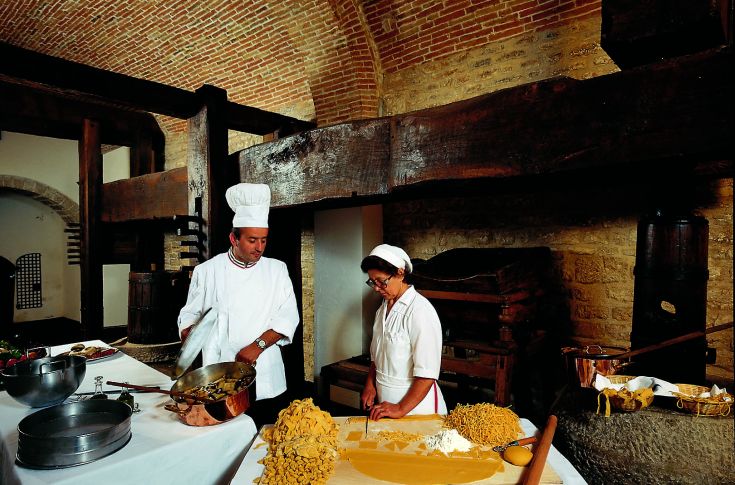 Deux chefs préparent des pâtes dans une cuisine rustique, entourés d'ustensiles traditionnels et de matériaux naturels.