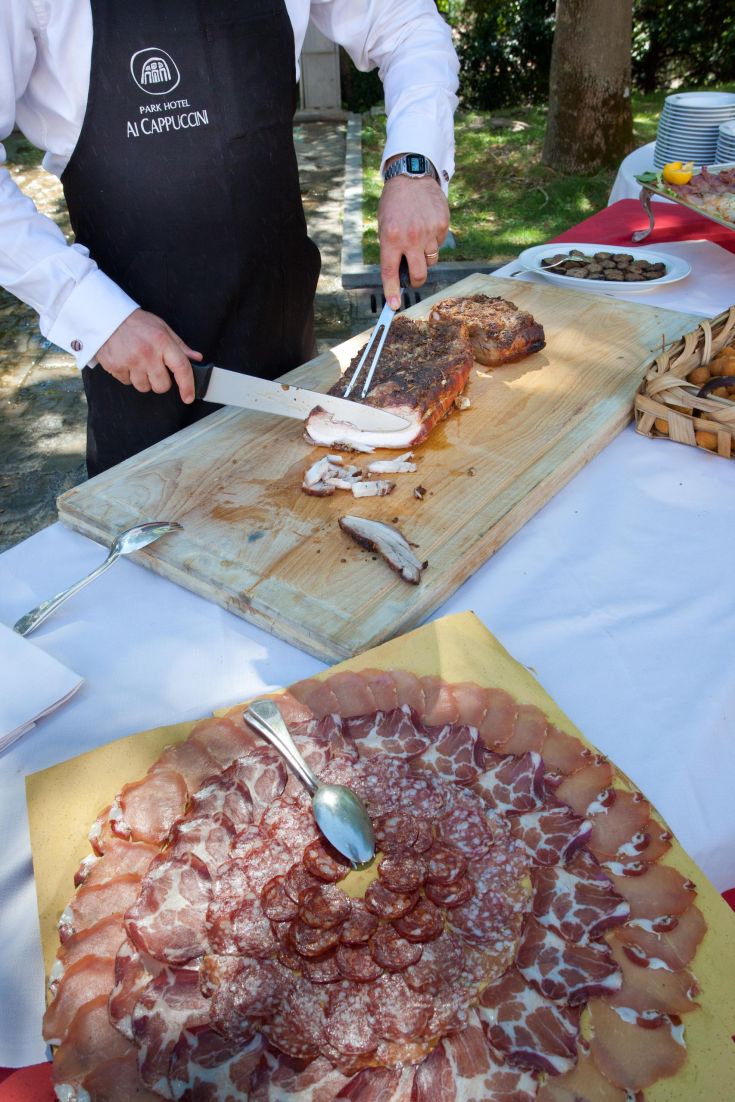 Un chef prépare des charcuteries sur une planche à découper, avec un assiette de tranches de charcuterie en évidence. Un cadre informel et accueillant.