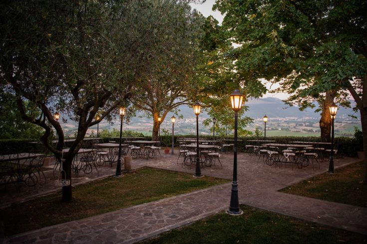 Ein einfacher Außenbereich mit Tischen, beleuchtet von Lampen und umgeben von Vegetation.