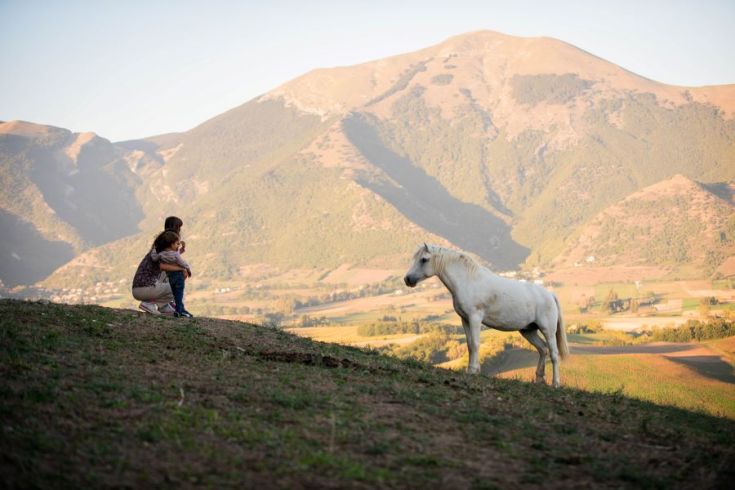 Ein junges Mädchen und ein Kind beobachten ein weißes Pferd in einer ruhigen Berglandschaft.