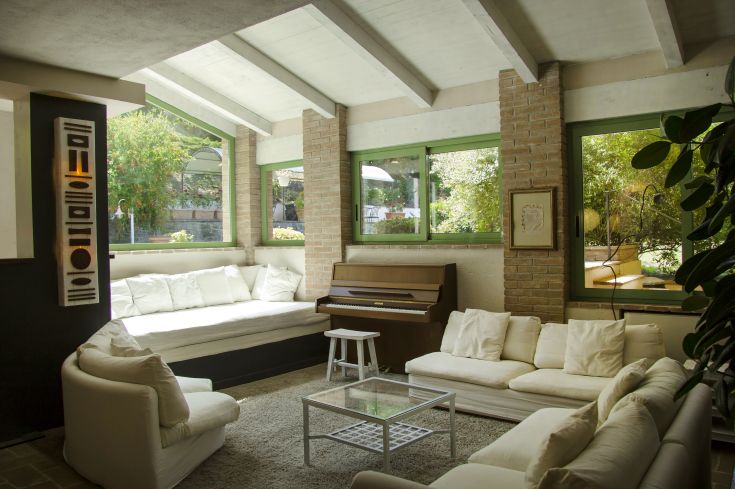 Stanza ben illuminata con divani bianchi e un pianoforte, circondata da un ambiente naturale.