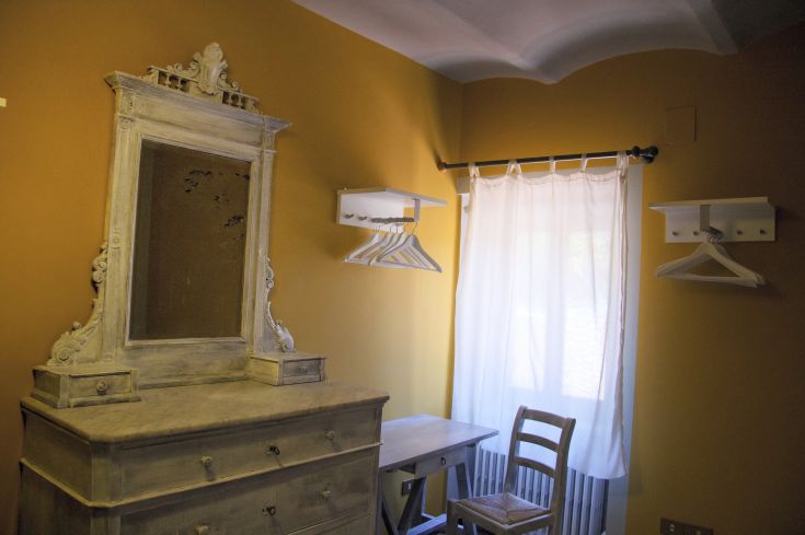 Interno di una stanza con arredamento sobrio, un mobile pratico, uno specchio e una finestra decorata da tende chiare.