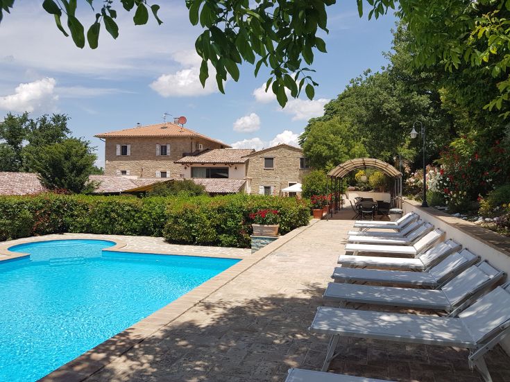 Una piscina di colore azzurro immersa in un giardino con piante e sedie a sdraio.