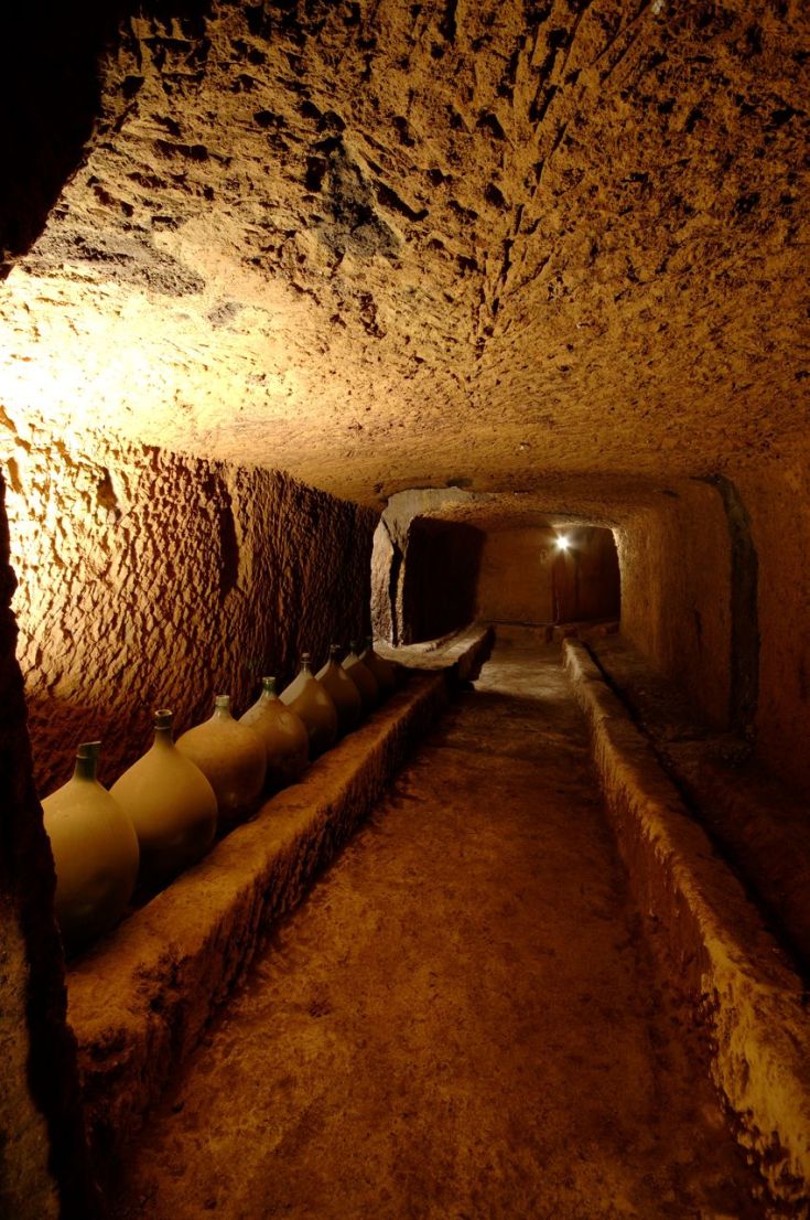 Una cantina storica situata nella roccia, con bottiglie di terracotta disposte su scaffali e illuminate da una luce tenue.