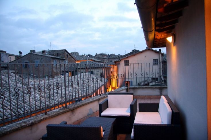 Scena di un terrazzo in un palazzo, arredato con elementi moderni e affacciato su tetti storici.