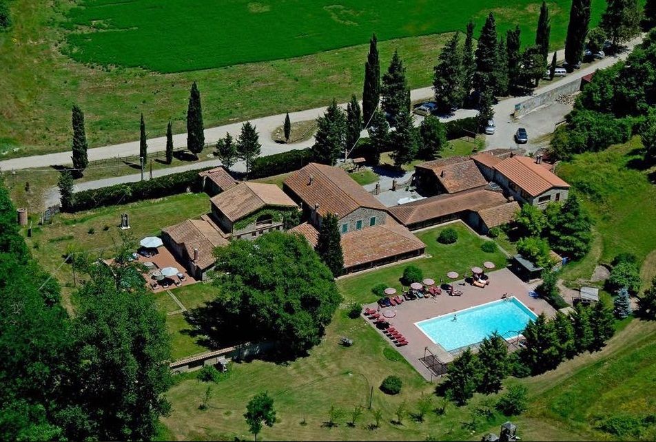 A view of an old farmhouse with a swimming pool, surrounded by vast green spaces.