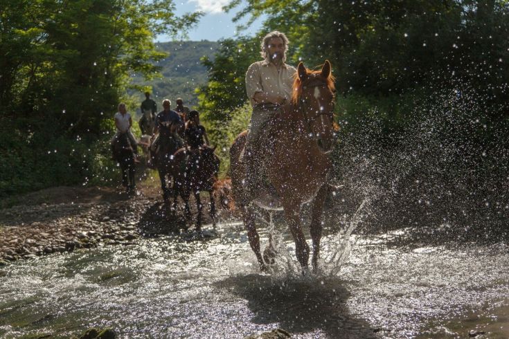 Ritter überqueren einen Bach und spritzen Wasser, während die Sonne über ihnen scheint.