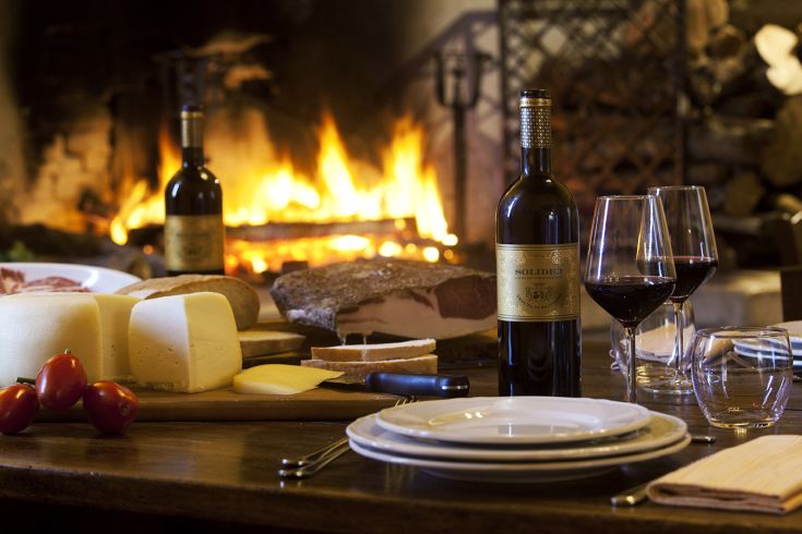 An intimate dining table set with wines, cheeses, and cold cuts, next to a lit fireplace.