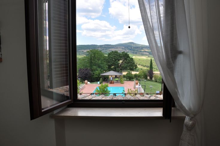 Scene through a large window of a farmhouse in Umbria, with a pool and green hills in the background.