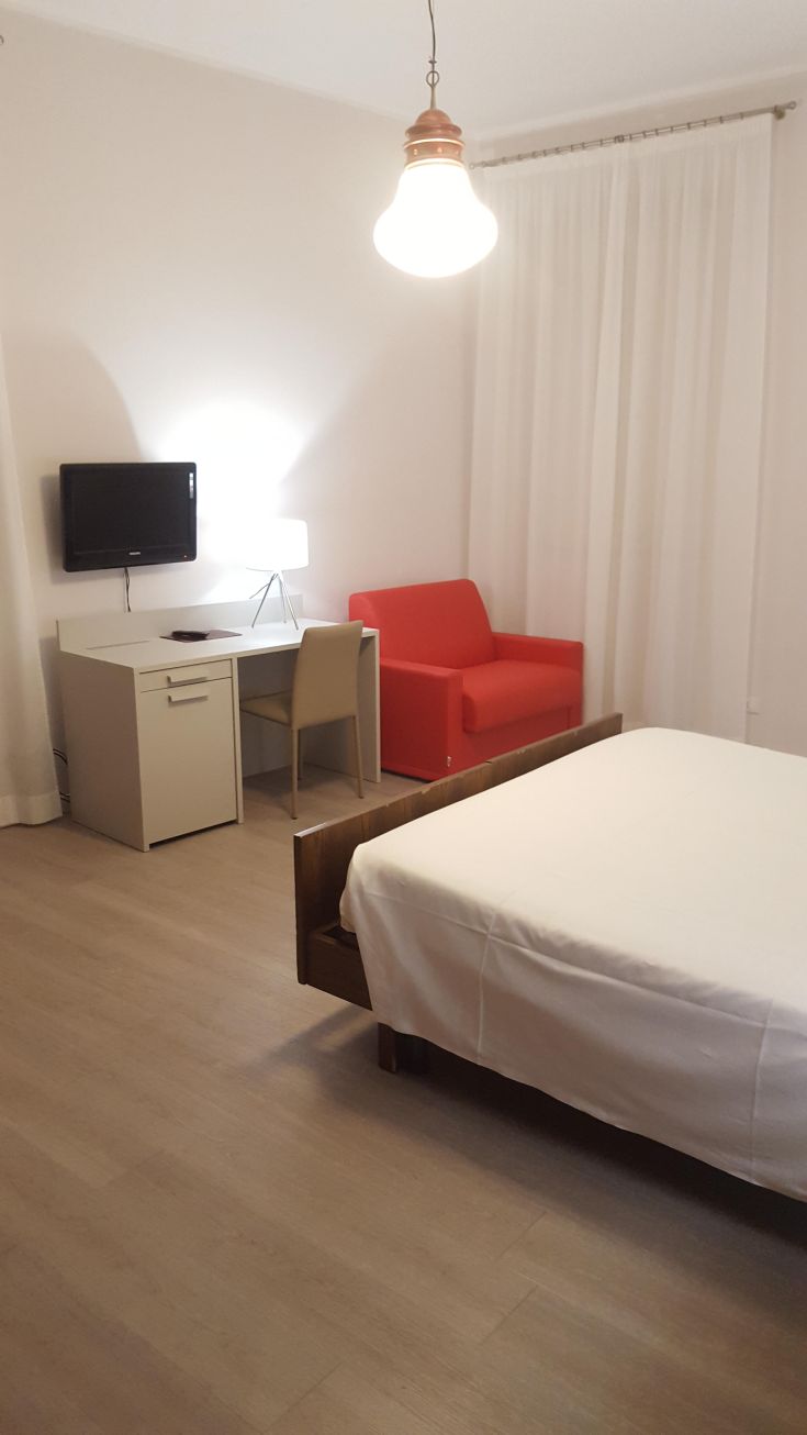Camera confortevole con mobili essenziali, completa di televisore, scrivania e un divano di colore rosso.