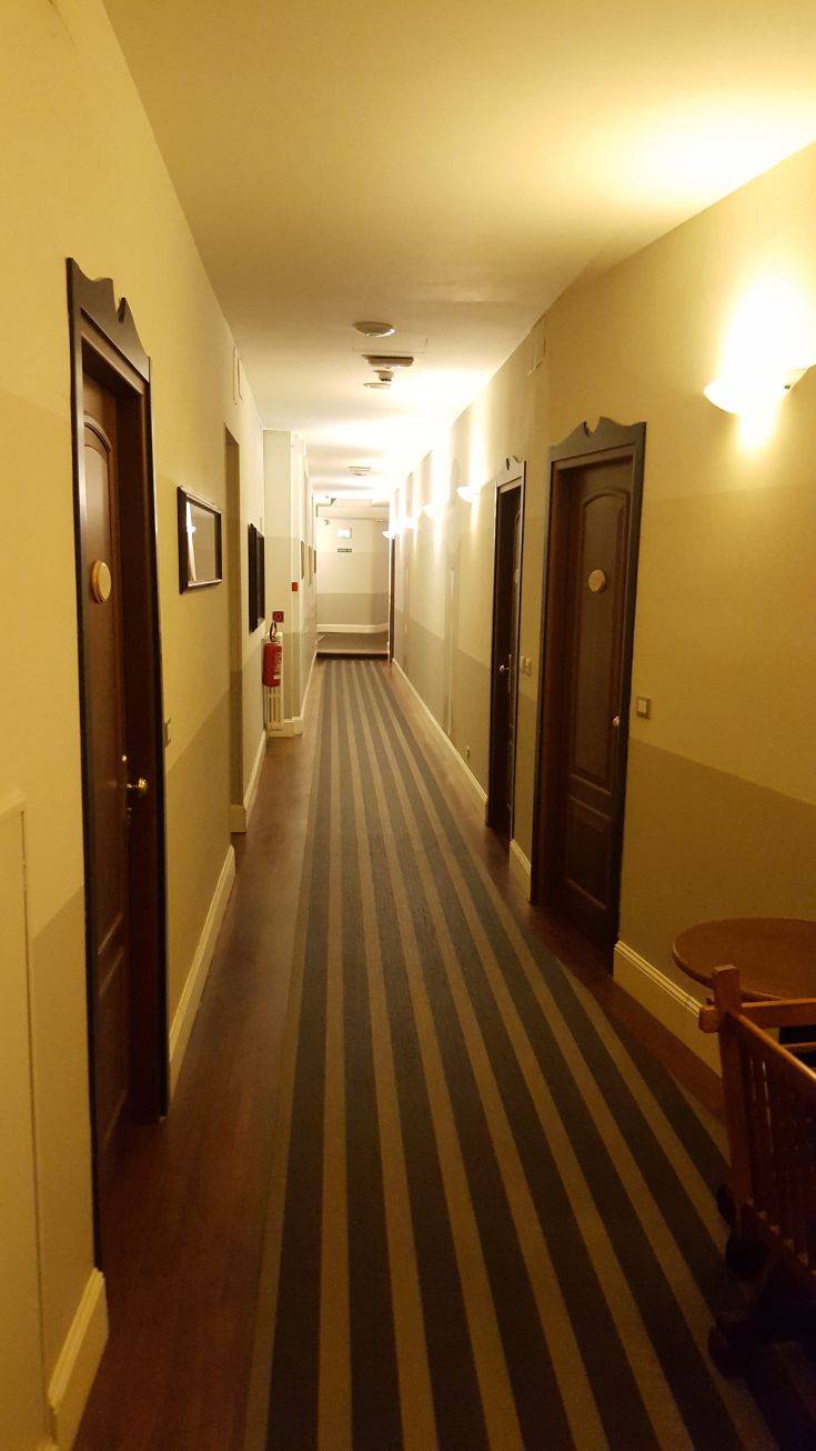 Corridoio luminoso di un hotel, con porte in legno e moquette a strisce. L'ambiente è caldo e caratteristico.