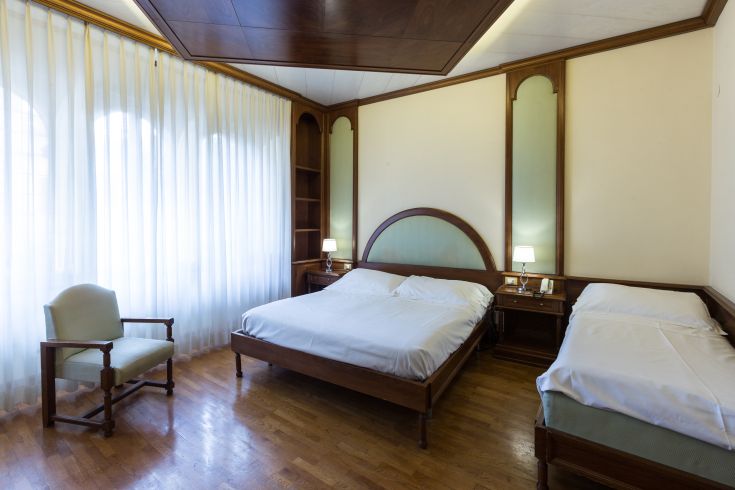 Camera semplice con letti singoli e matrimoniali, illuminata da tende chiare e arredo in legno.