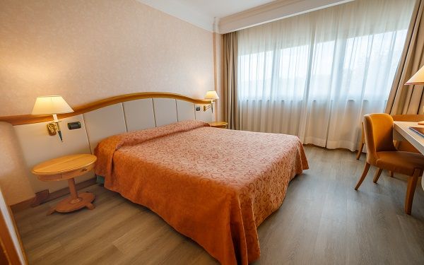 A bright and simple hotel room featuring a double bed and a desk. The window provides ample natural light.