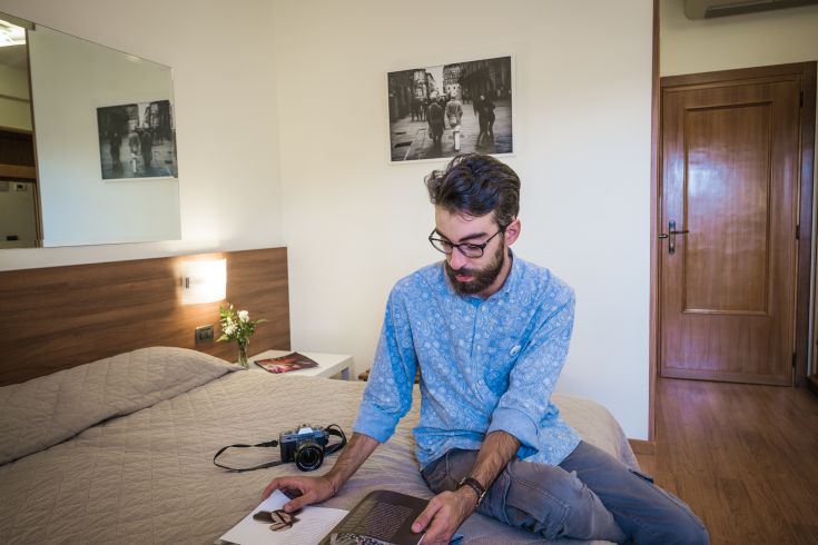 Ein junger Mann blättert in einem Buch in einem einfachen Hotelzimmer mit funktionalem Dekor.