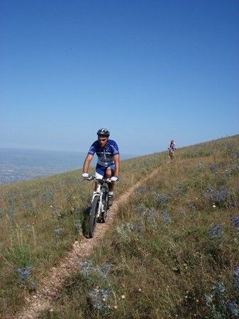 A cyclist rides on a mountain trail, surrounded by lush vegetation and under a sunny sky.
