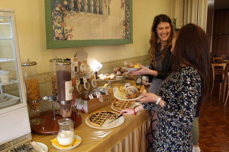 Two friends smile while enjoying a breakfast buffet filled with pastries and fresh dishes.