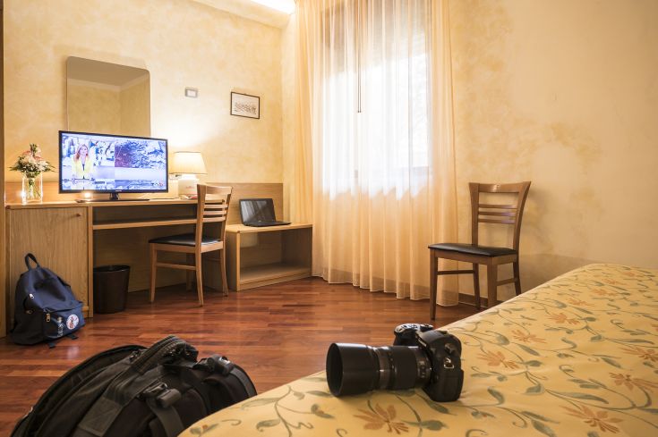 Simple and comfortable hotel room featuring a TV, desk, and bed. Natural light floods in through the window with curtains.