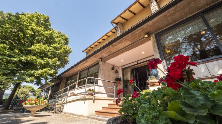 A hotel located in a green area, featuring a garden with flowers and an outdoor swimming pool.