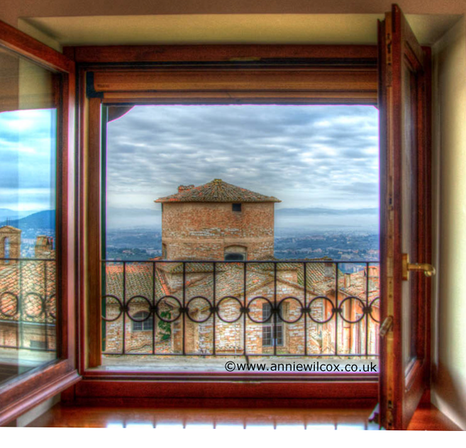 Scena dall'interno di una finestra che mostra il panorama sui tetti e sulla torre di un centro storico dell'Umbria.