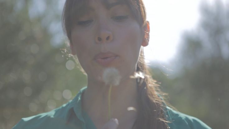 Eine Frau bläst auf einen Löwenzahn in einer ruhigen und hellen Naturlandschaft.