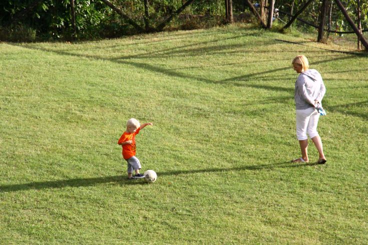 Ein Kind tritt einen Ball auf dem Rasen, während eine Frau aufmerksam zusieht.