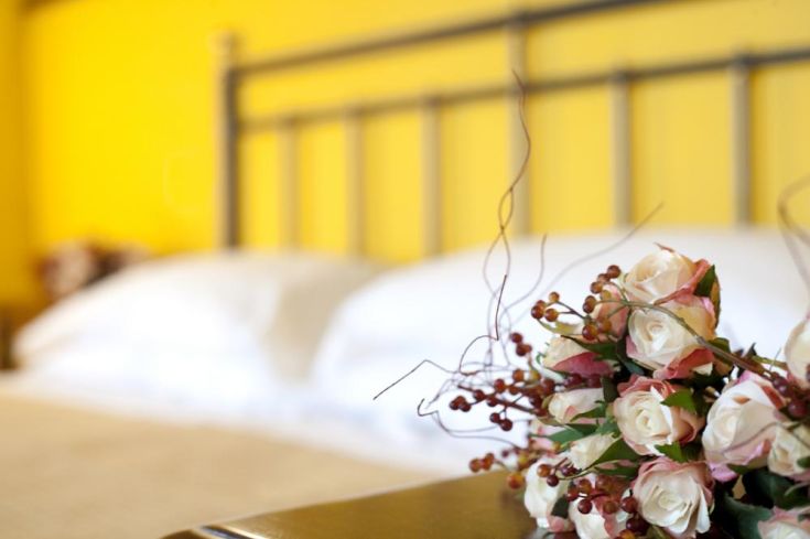 A bouquet of flowers placed next to a bed with white linens in a room with yellow walls.